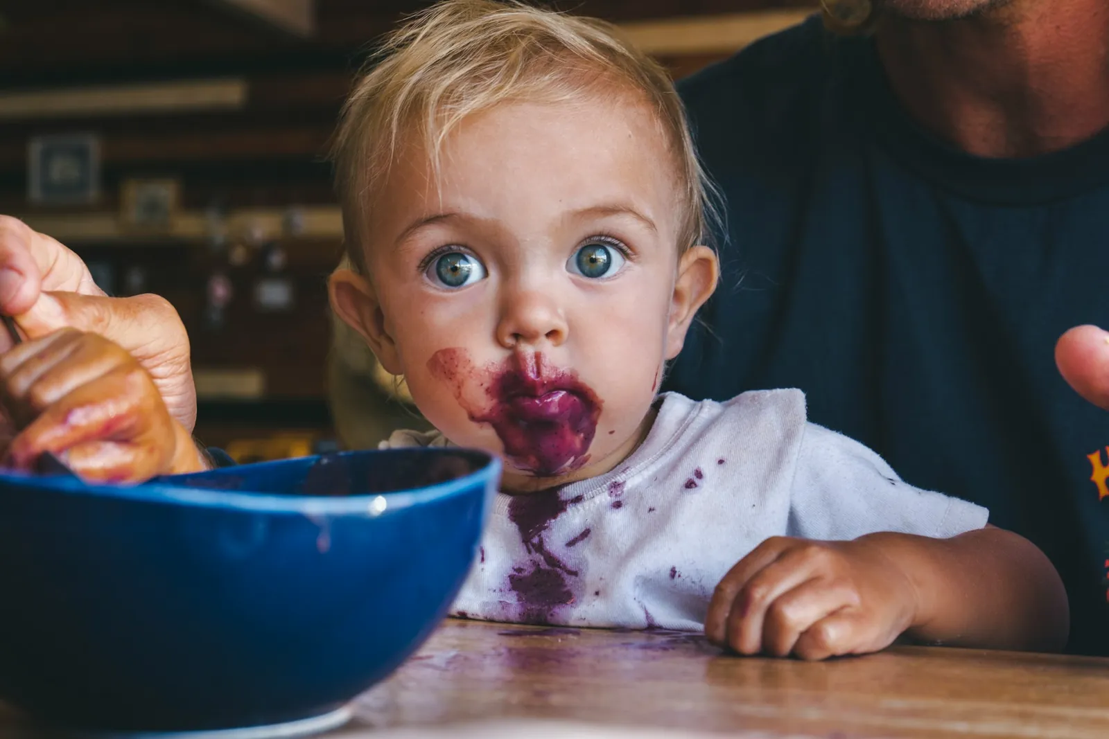 Ein Kleinkind mit großen Augen wird aus einer blauen Schüssel gefüttert, Gesicht und Shirt voller violettem Beerenbrei